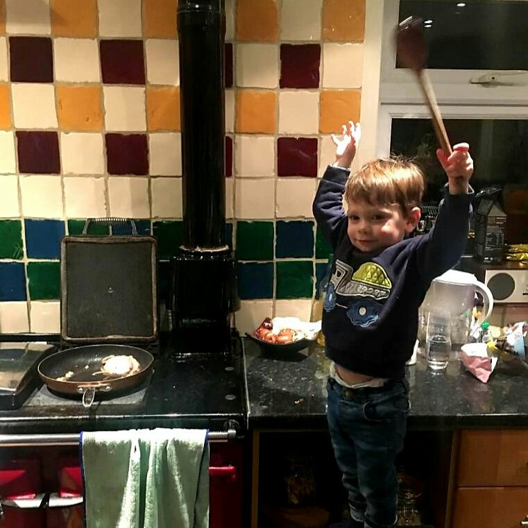 Young boy in a kitchen, holding a wooden spoon up, next to a stove with a frying pan.