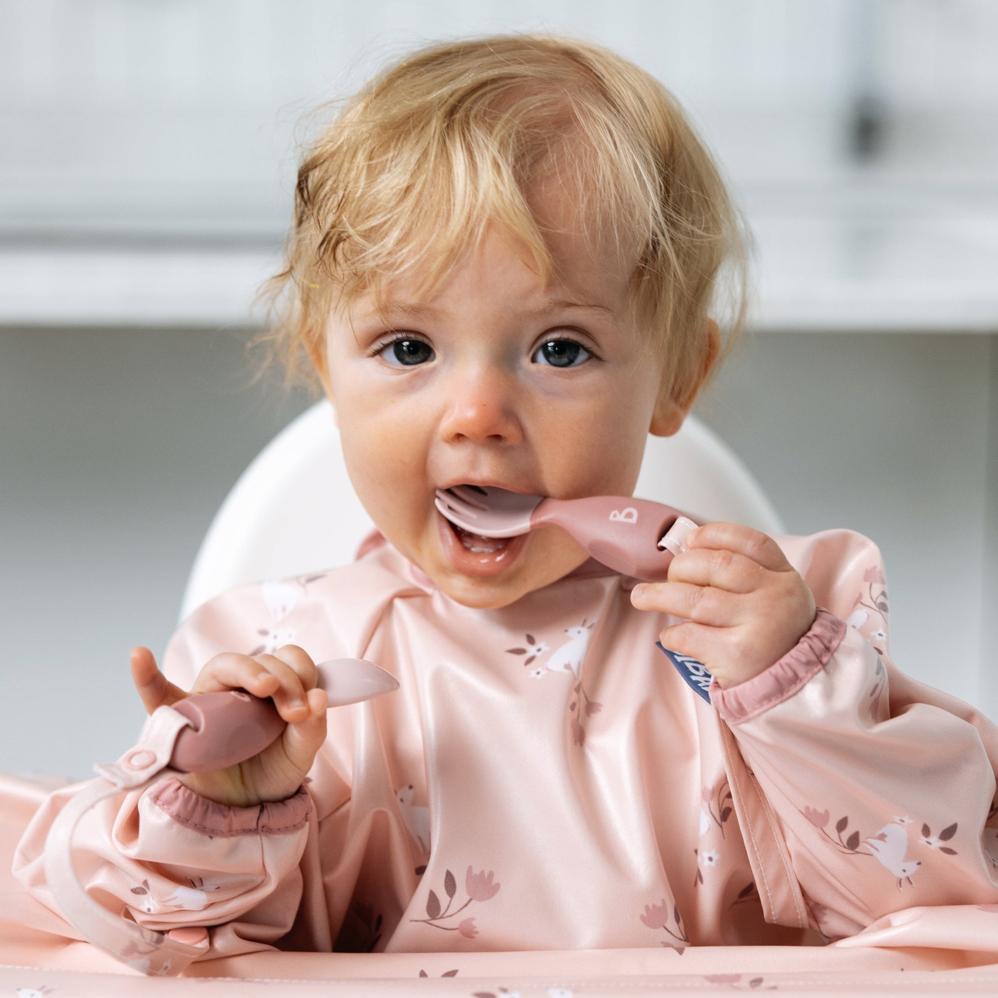 Blonde baby in a pink bib, holding a pink silicone spoon to their mouth.