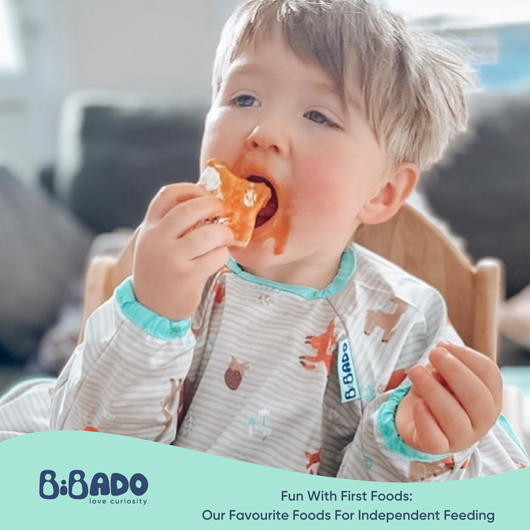 Toddler in a patterned bib eating a messy piece of food independently.