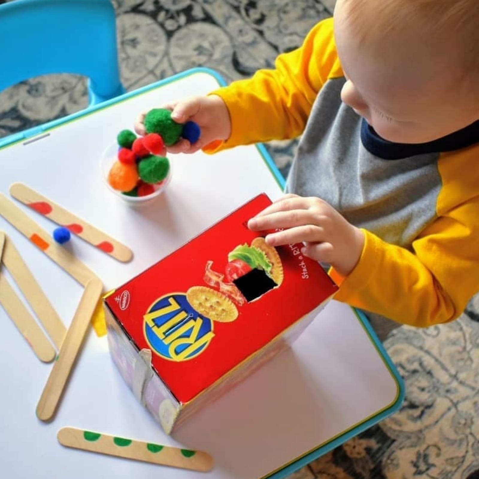 Toddler playing with a red Ritz cracker box, pushing colourful pom-poms into a cut-out hole.