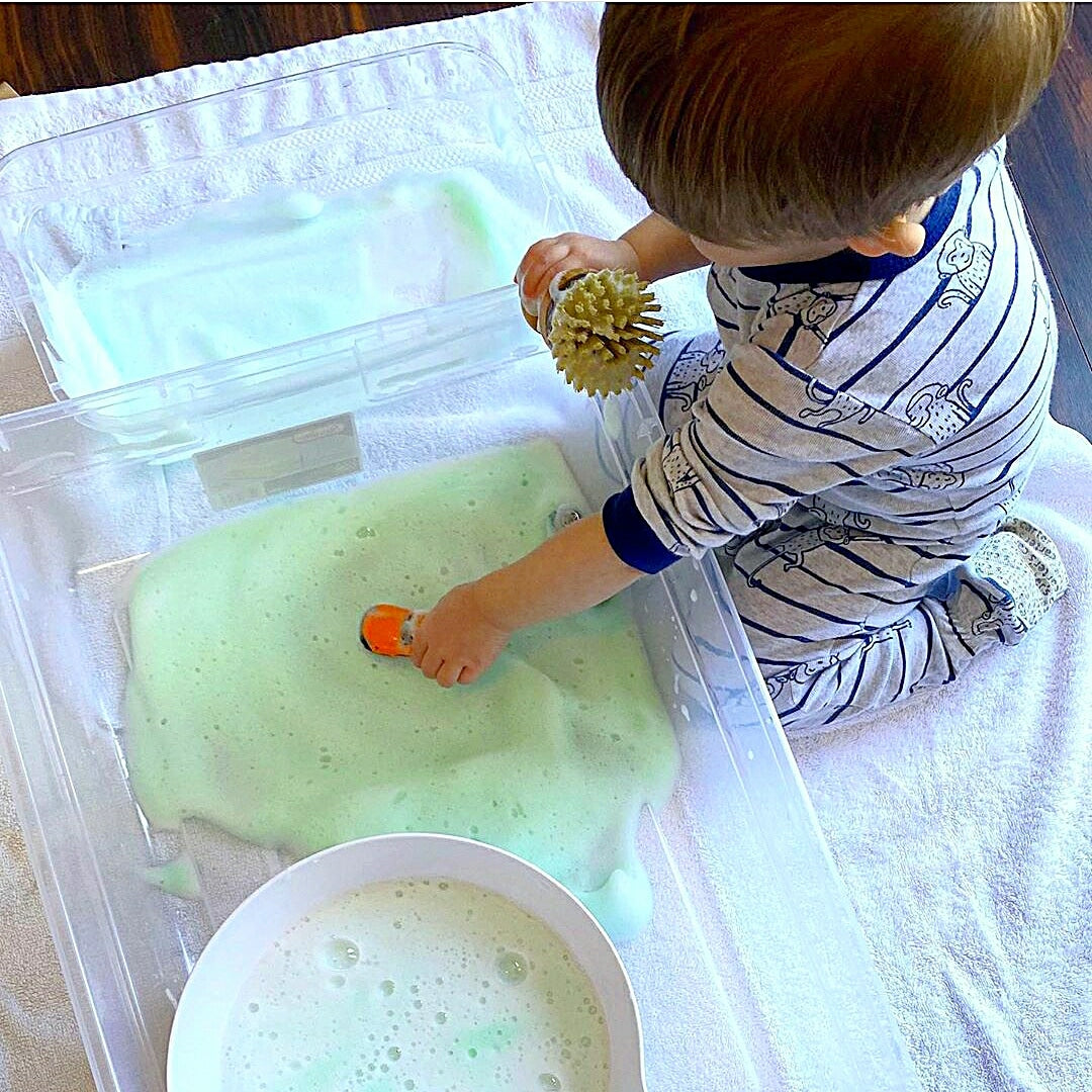 Toddler playing with green bubble foam and toy car in clear tubs on a white towel.