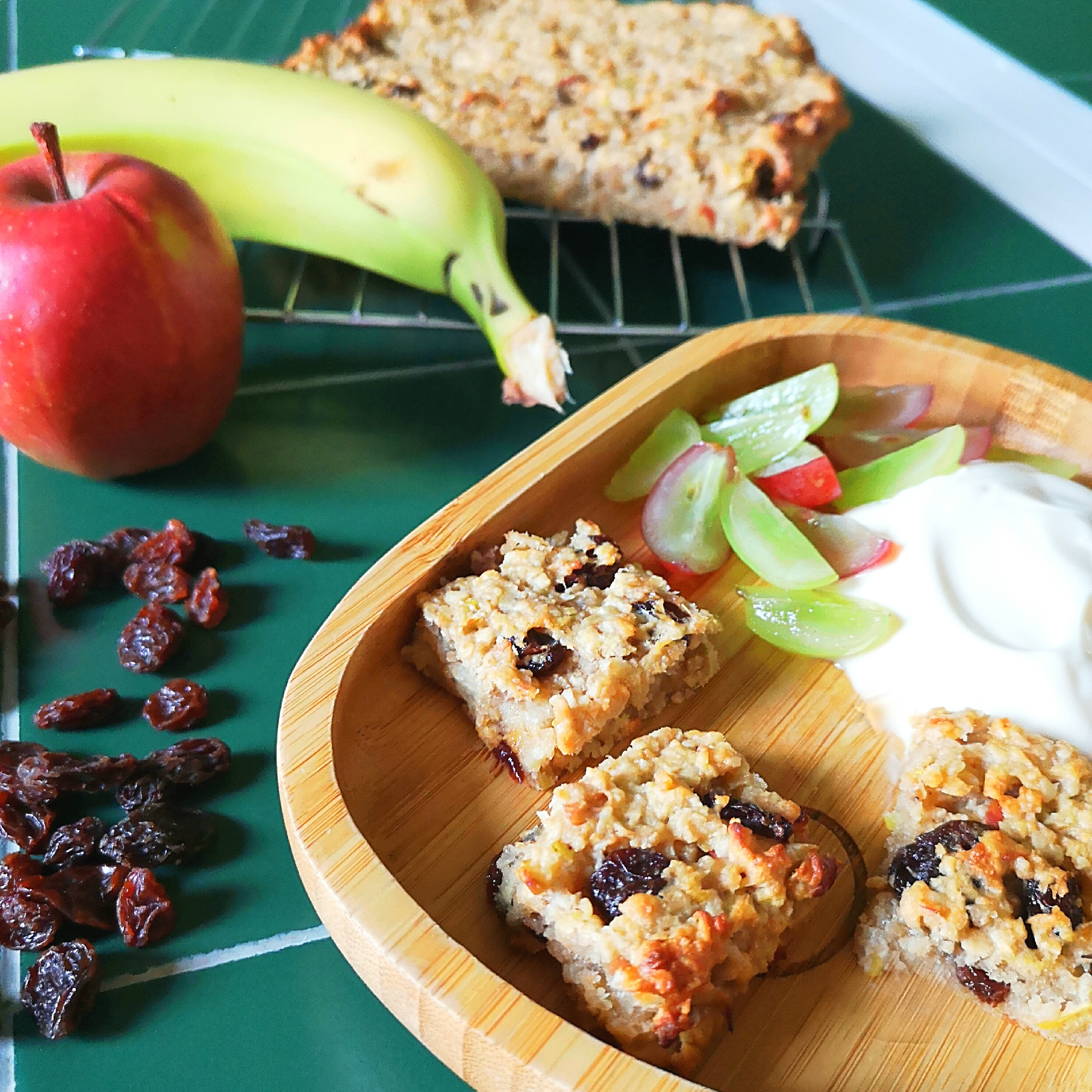 Homemade fruit & oat bars with raisins, banana, apple, grapes, and yoghurt on a bamboo plate.