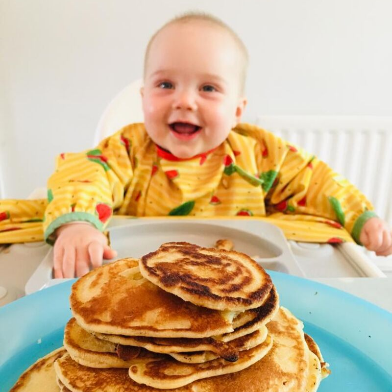 Smiling baby in a yellow bib, ready for a stack of pancakes on a blue plate.