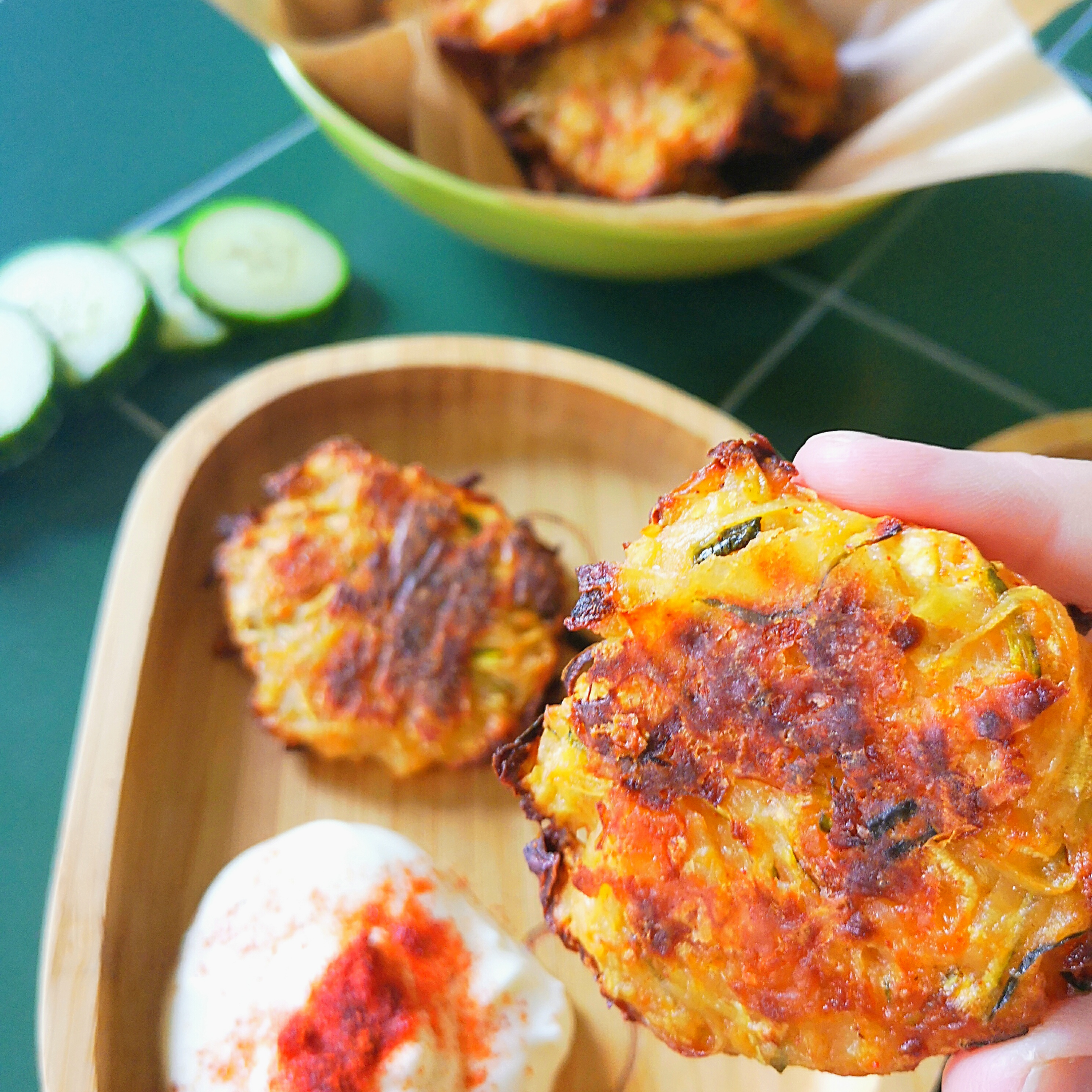 Hand holding a golden-brown courgette patty over a wooden plate with dip and cucumber slices.