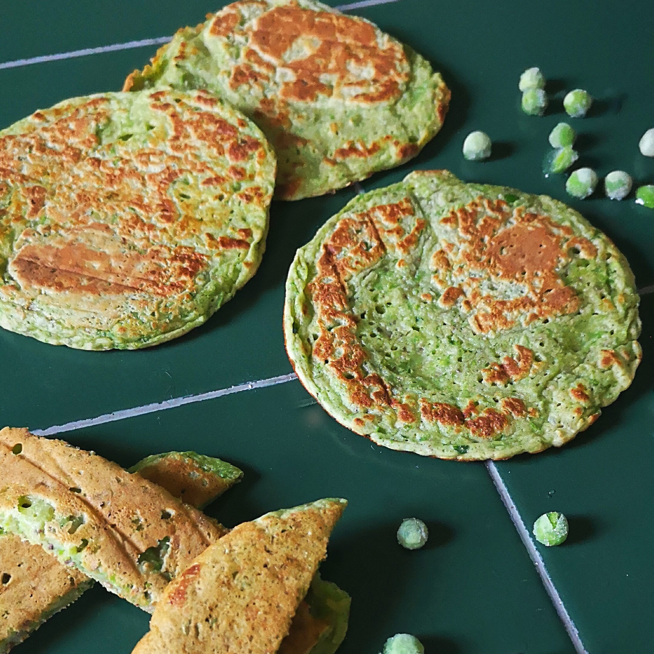 Golden-brown pea pancakes with vibrant green peas scattered on a dark green surface.