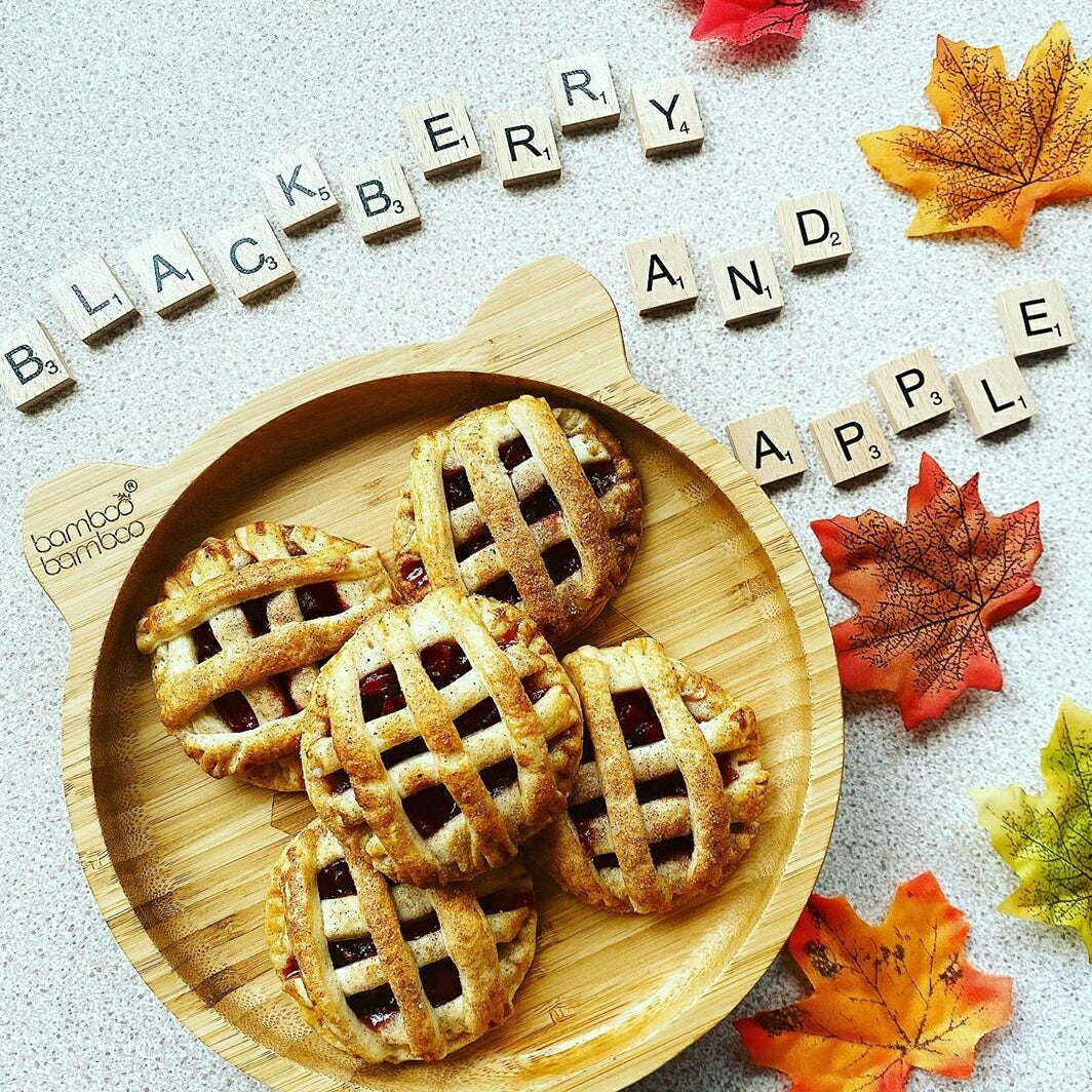 Five mini lattice-top apple and blackberry pies on a bamboo plate with autumn leaves and Scrabble tiles.