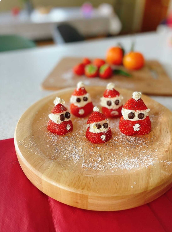 Five festive strawberry Santas with cream beards and chocolate chip eyes on a wooden plate.
