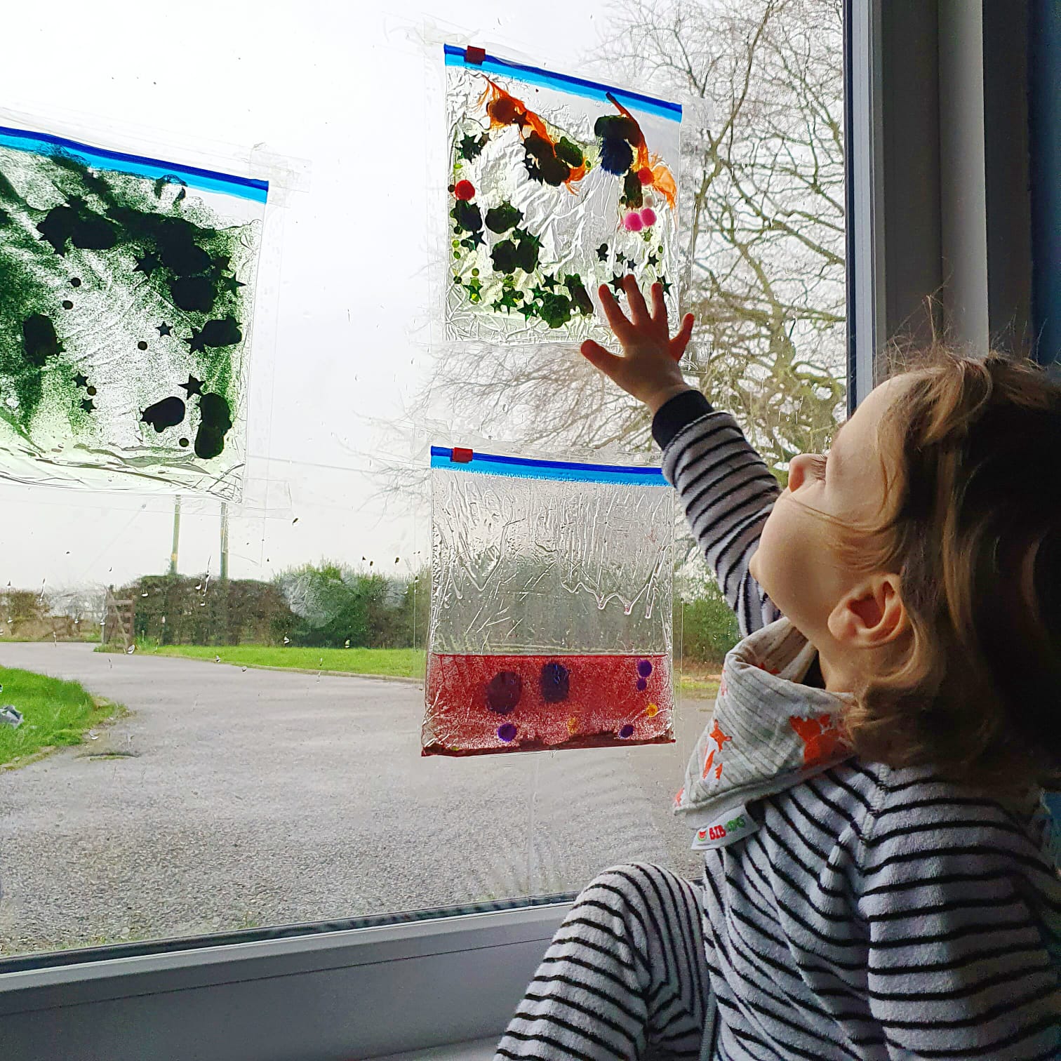 Toddler reaching for colourful sensory bags taped to a window.