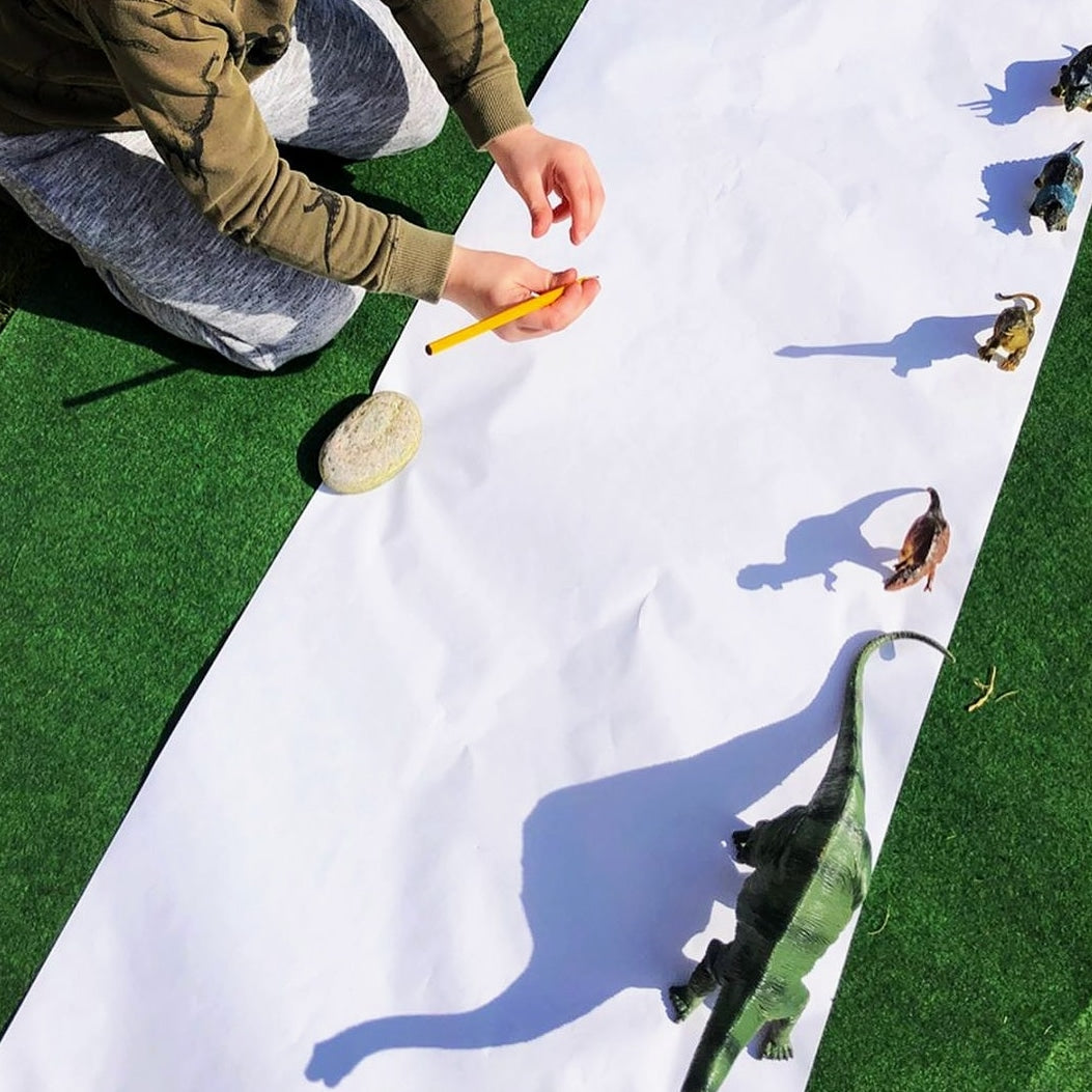 Child drawing dinosaur shadows on a long white paper roll outdoors.