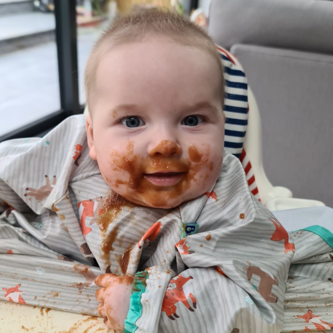 Messy baby in a highchair, covered in food, smiling at the camera.