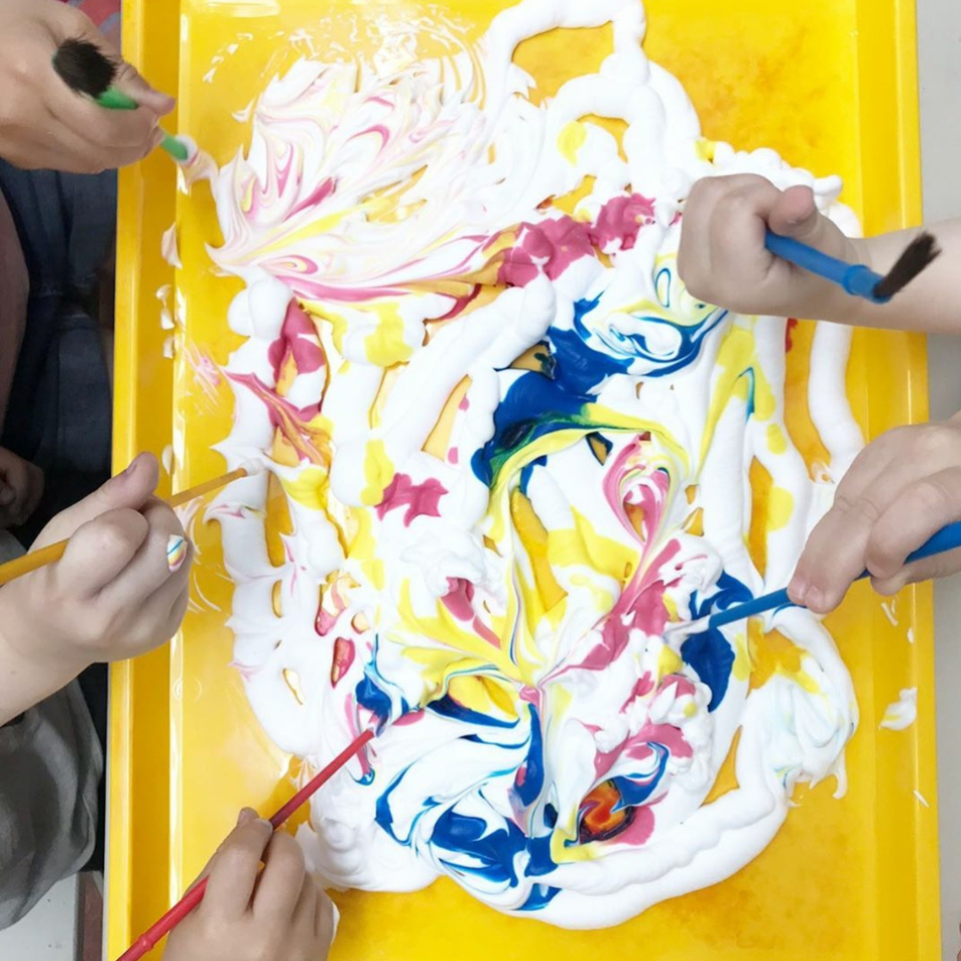 Children's hands painting vibrant colours into white shaving foam on a yellow tray.