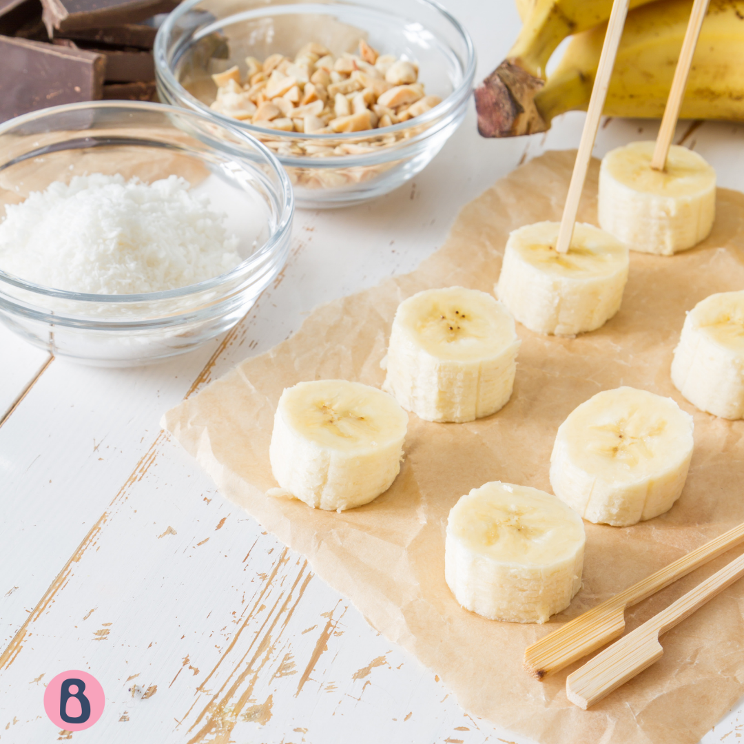 Banana slices on sticks with bowls of shredded coconut, chopped nuts, and chocolate.