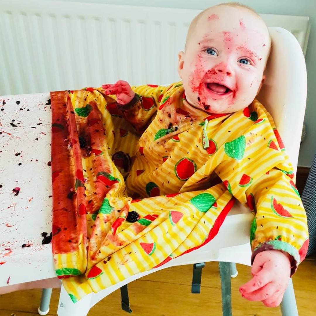 Happy baby in a highchair, covered in berry puree from baby-led weaning.