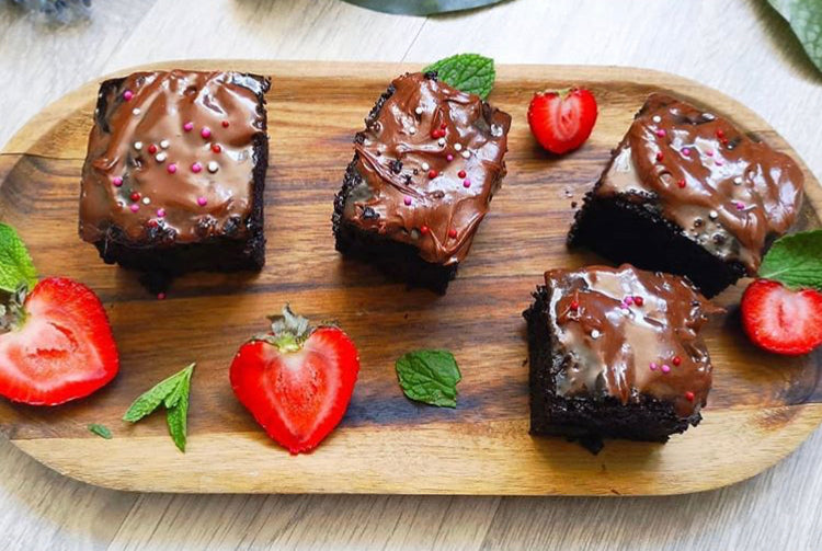 Four rich courgette brownies with chocolate icing and sprinkles, garnished with strawberries and mint on a wooden board.