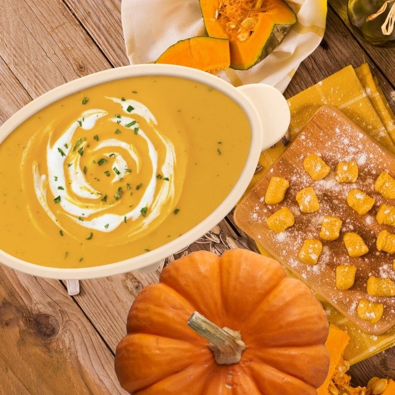 Creamy pumpkin soup in a white bowl, garnished with cream and parsley, beside a whole pumpkin and gnocchi.