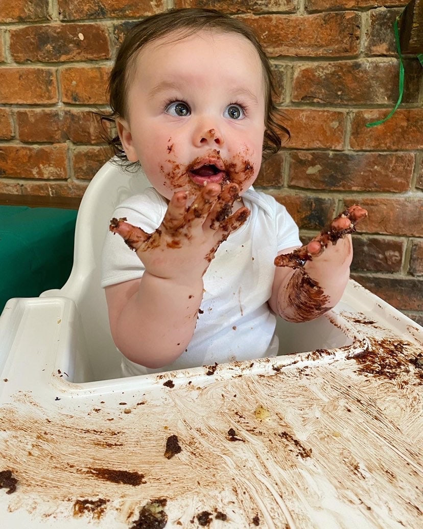 Messy baby in a white highchair, covered in chocolate cake, looking up.