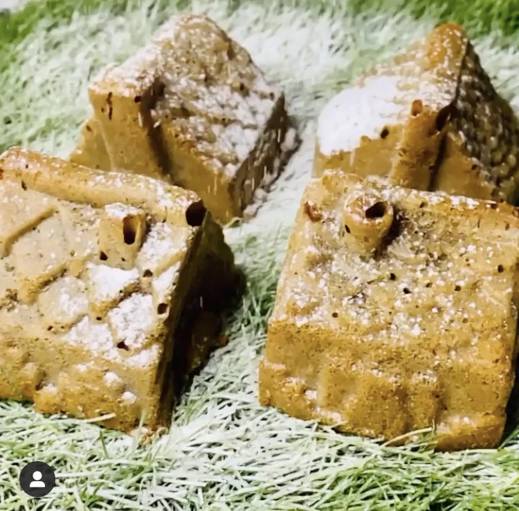 Four golden-brown carrot and banana muffins shaped like houses, dusted with icing sugar on green.
