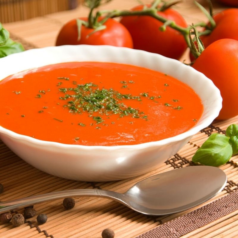 Vibrant red teething soup in a white bowl, garnished with herbs, surrounded by fresh tomatoes and basil.