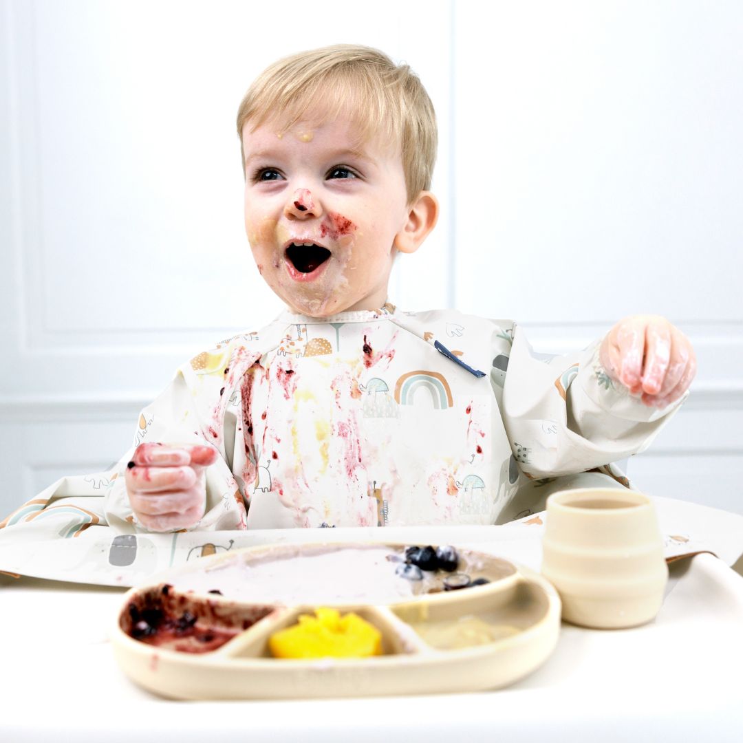 Messy blonde toddler in a patterned bib, covered in pureed food, enjoying baby-led weaning.