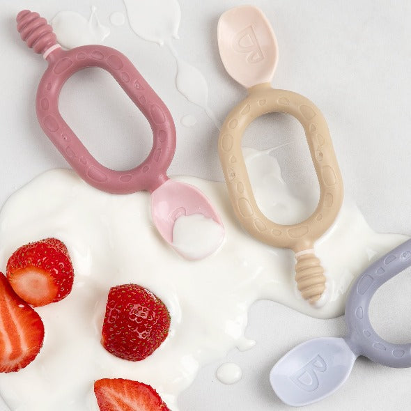 Two Dippit™ multi-stage baby spoons in pink and beige, with textured dippers and easy-grip handles, on spilled yogurt with strawberries. - Product image