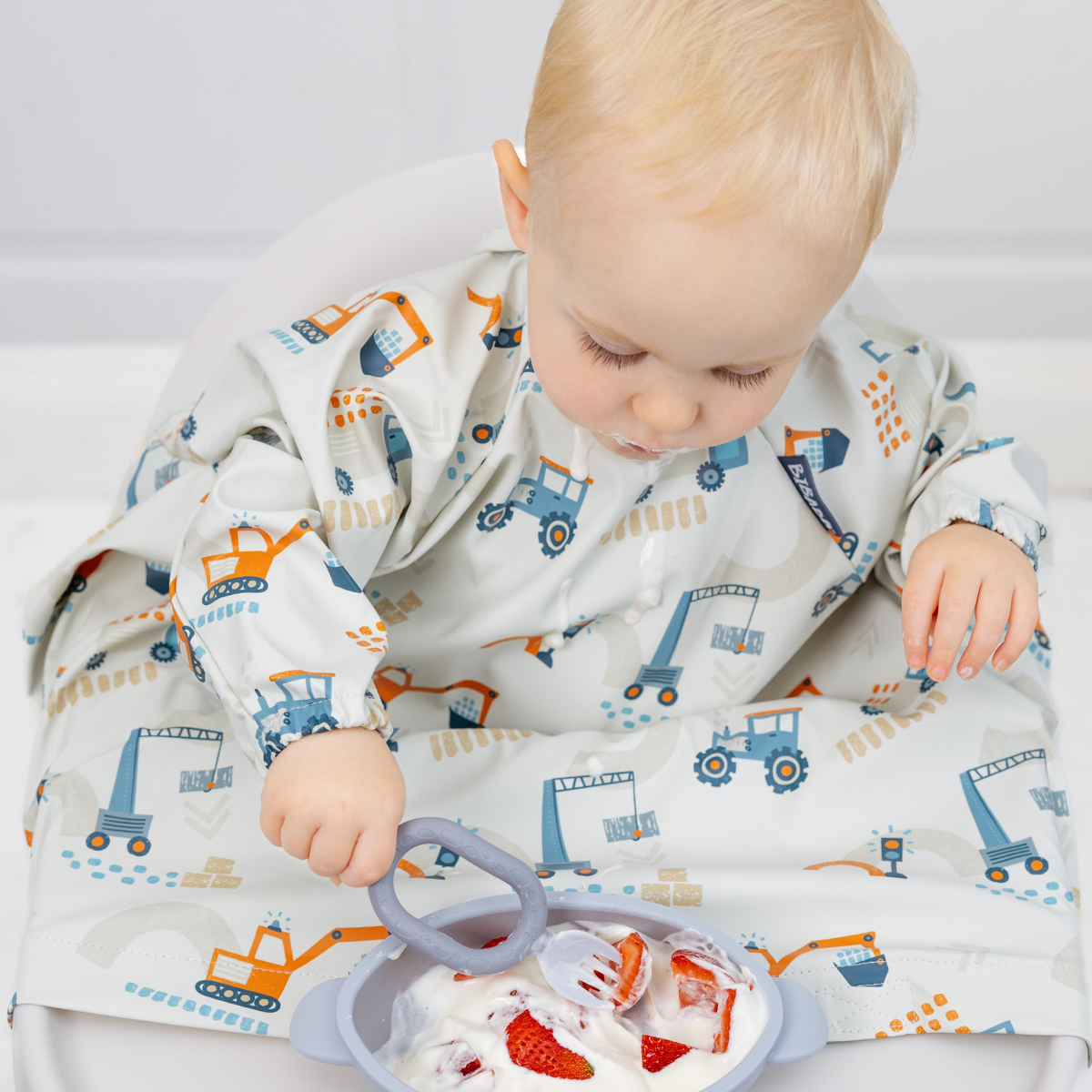 Toddler in a long-sleeved, light gray construction vehicle bib, holding a gray silicone spoon over a bowl of strawberries and yogurt. - Product image