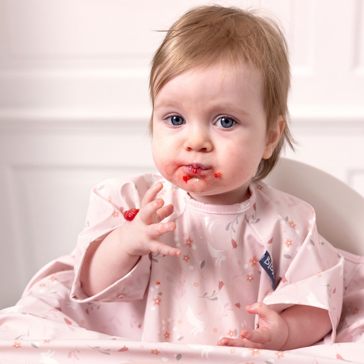 Baby in a pink short-sleeve coverall weaning bib with floral and unicorn print, messy from eating berries. - Product image