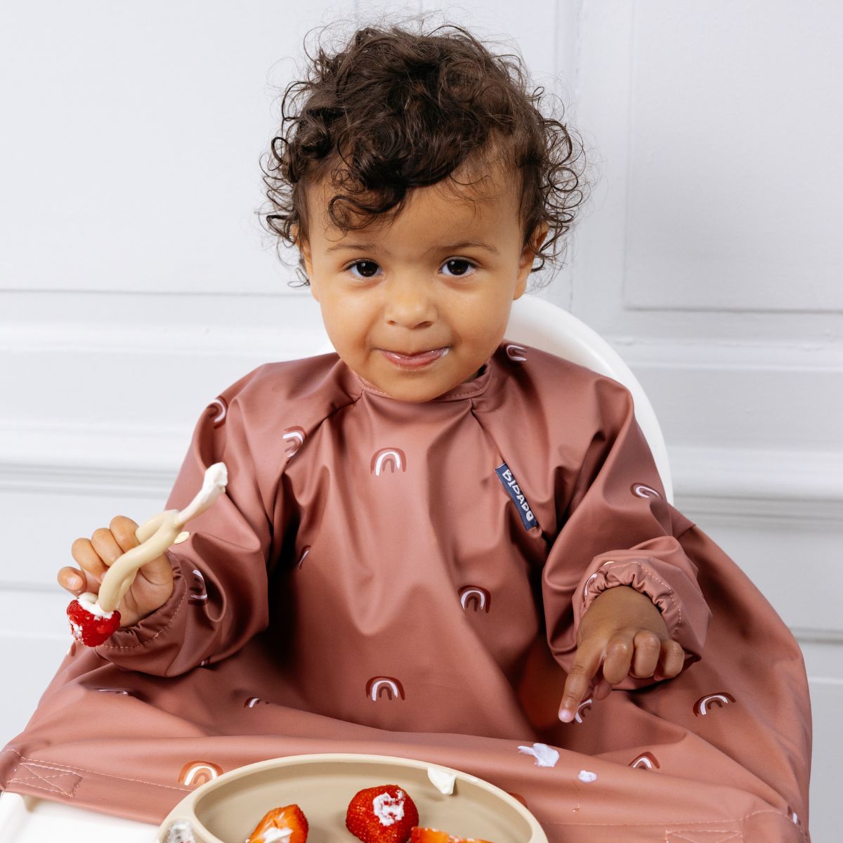 Toddler in a long-sleeve, dusty pink weaning bib with rainbow print, eating strawberries. - Product image