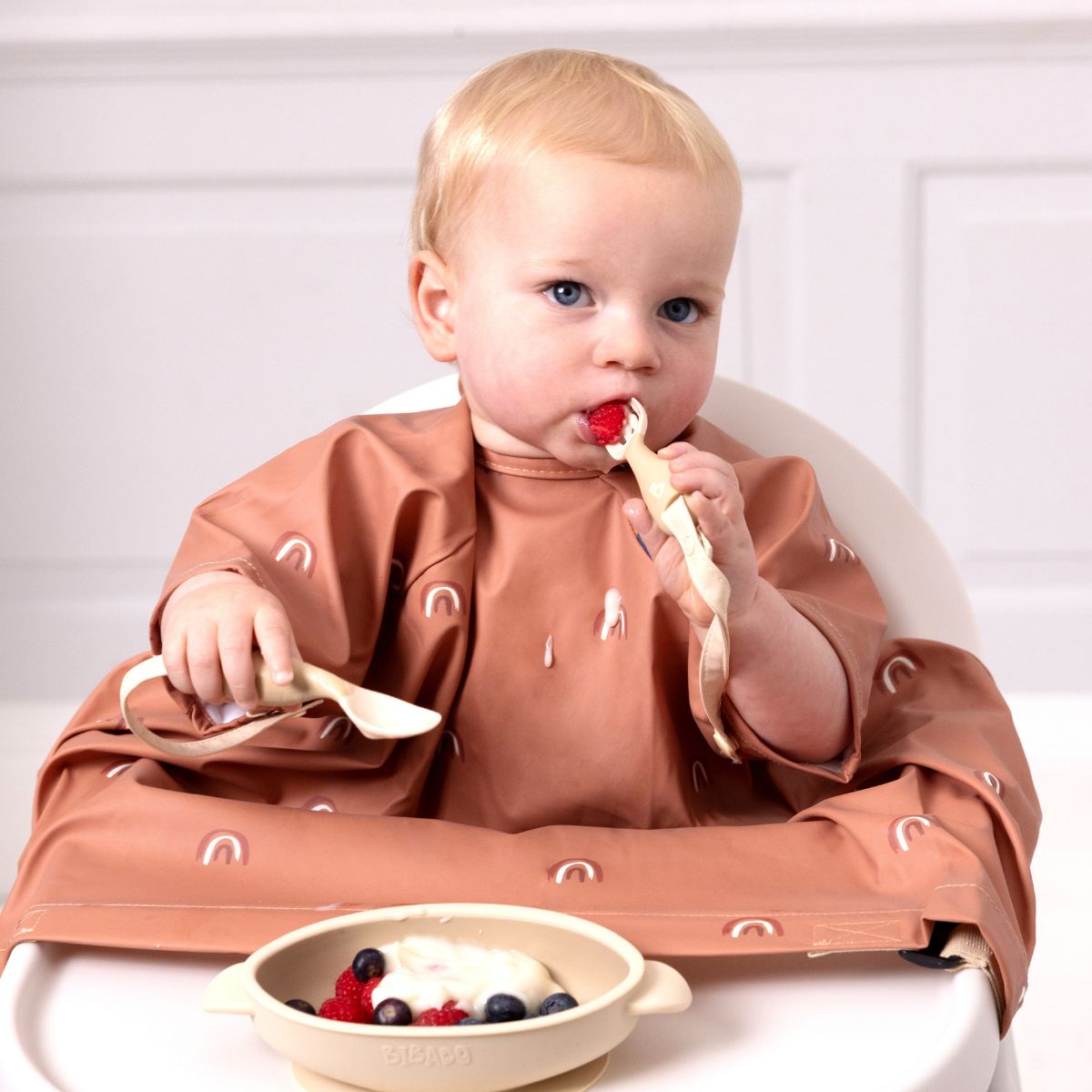 Baby in a short-sleeve terracotta Bibado coverall weaning bib with rainbow print, self-feeding with a spoon. - Product image