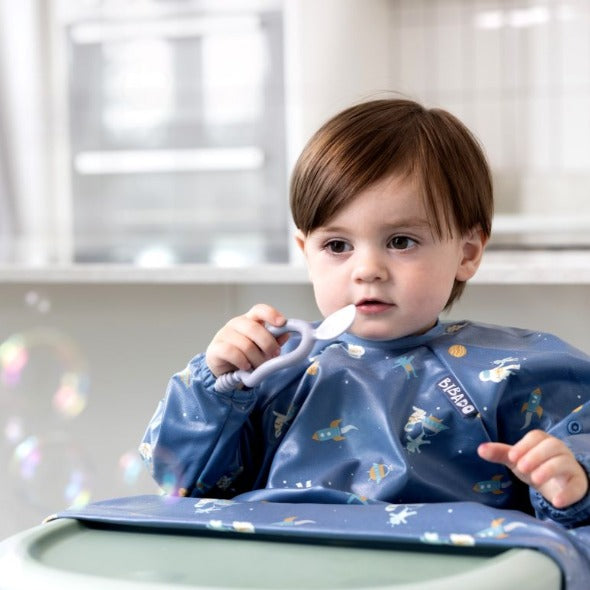 Baby in a blue space-themed bib holding a white Dippit™ multi-stage spoon with a textured dipper end. - Product image