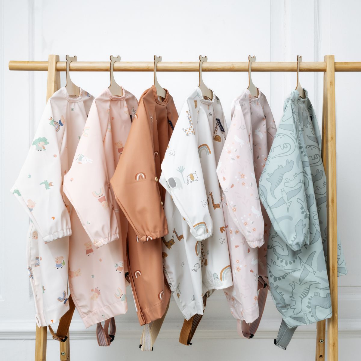 Six long-sleeved baby bibs in various pastel colors and animal prints hang on a wooden rack. - Product image