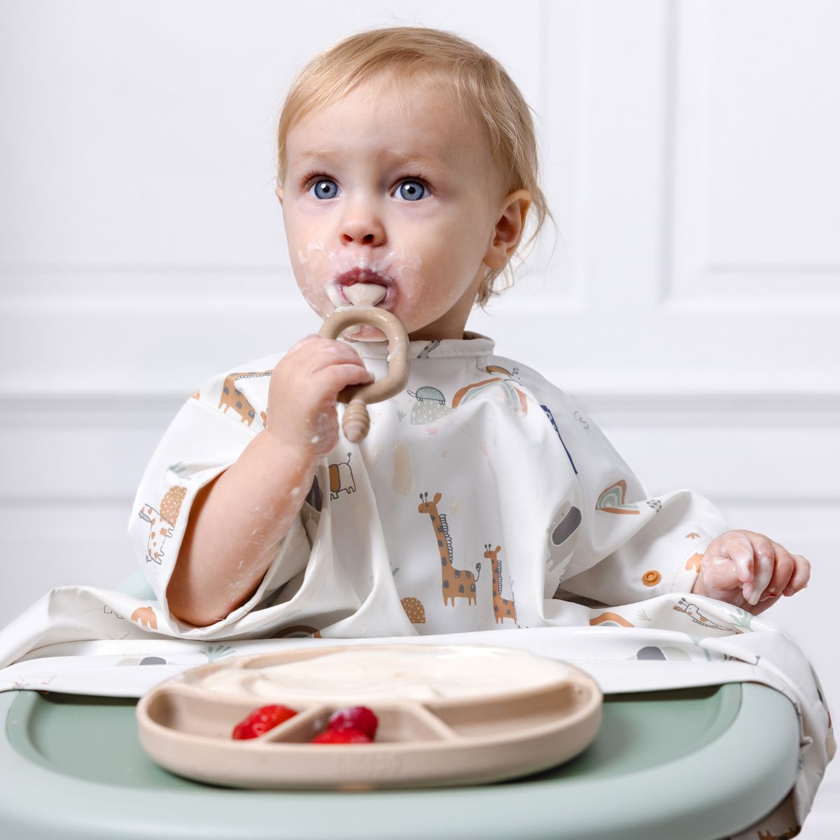 Blond baby in a long-sleeve white coverall weaning bib with giraffe print, eating from a beige plate. - Product image