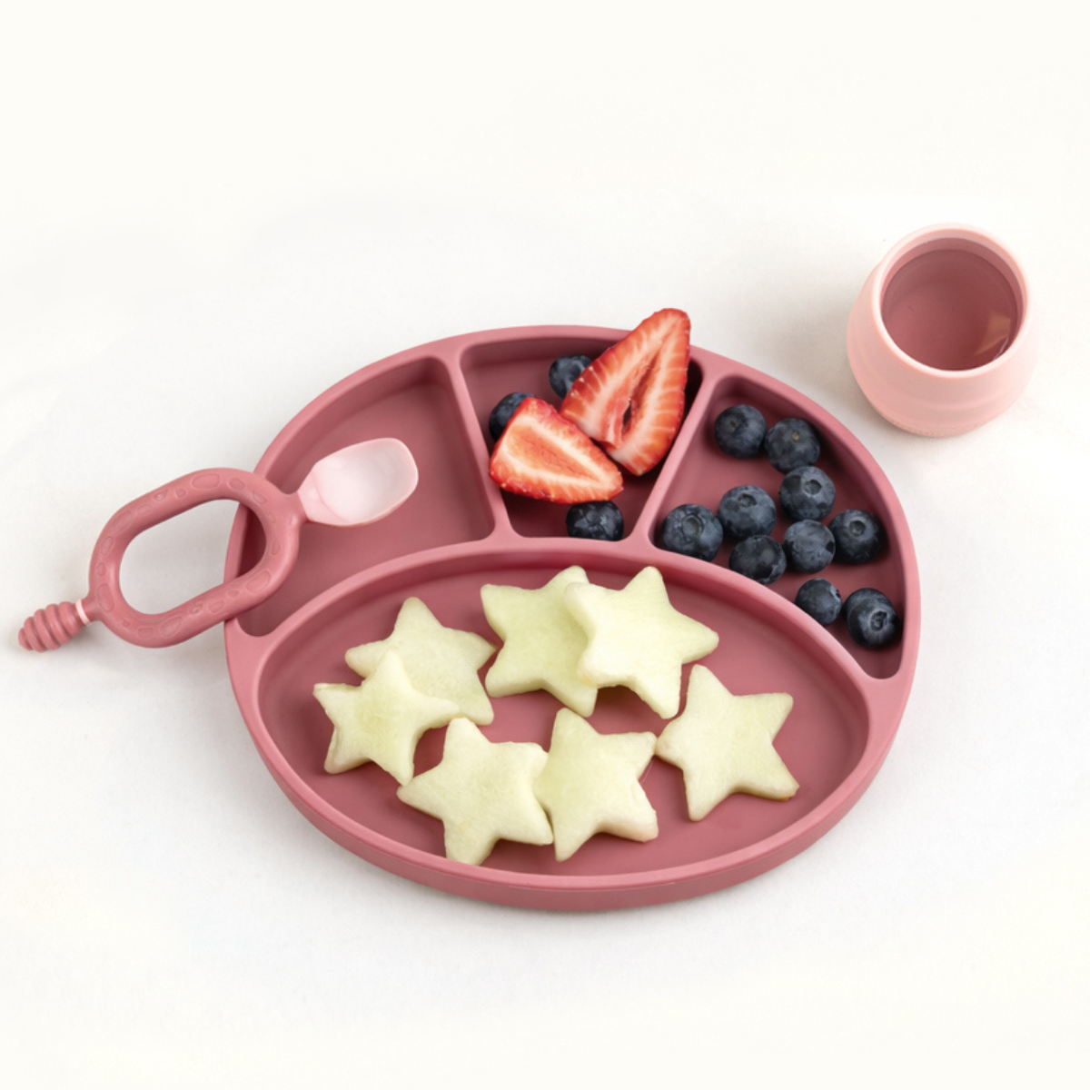 Pink /blush silicone plate with compartments filled with star-shaped fruit on a white background - Product image