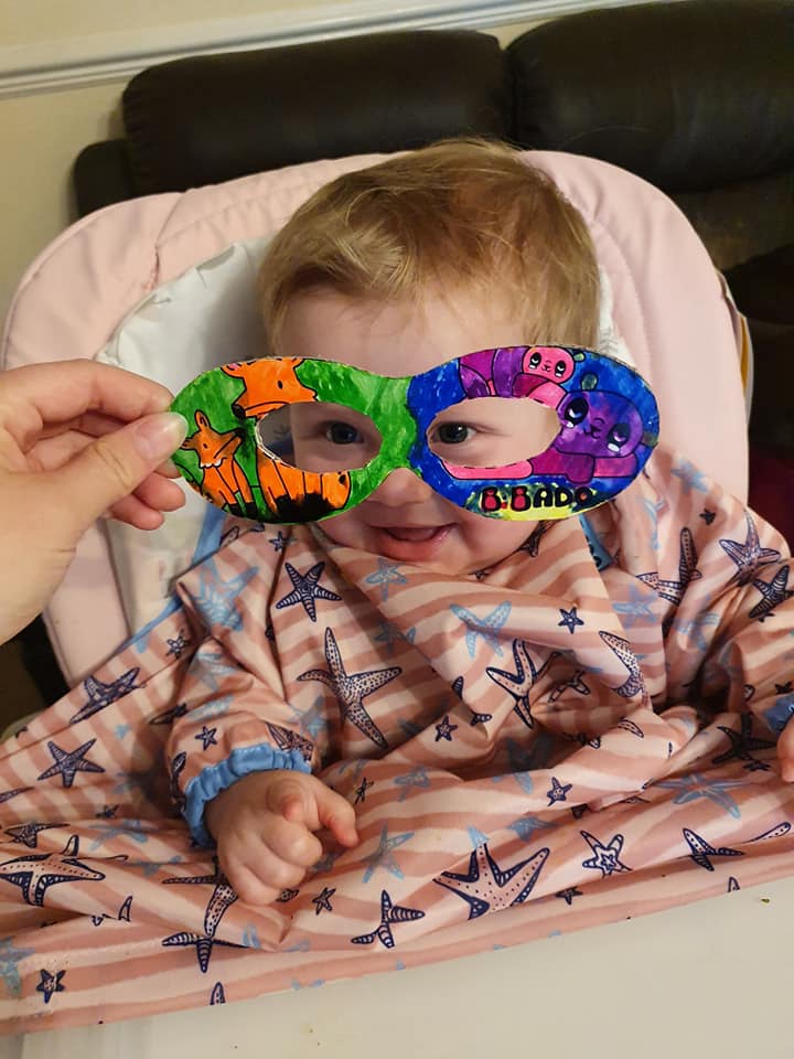 Baby in a high chair wearing a colourful, hand-drawn superhero mask.