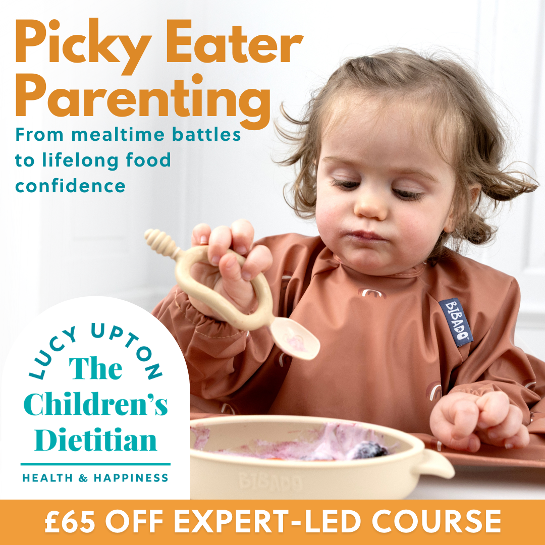 Toddler in a brown bib holding a spoon, looking at a bowl of food.