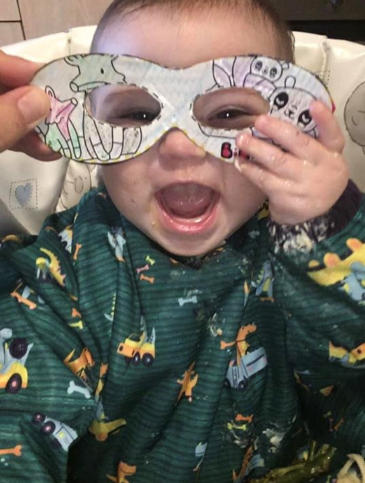 Messy baby in a green bib, laughing with a colourful drawn mask over their eyes.