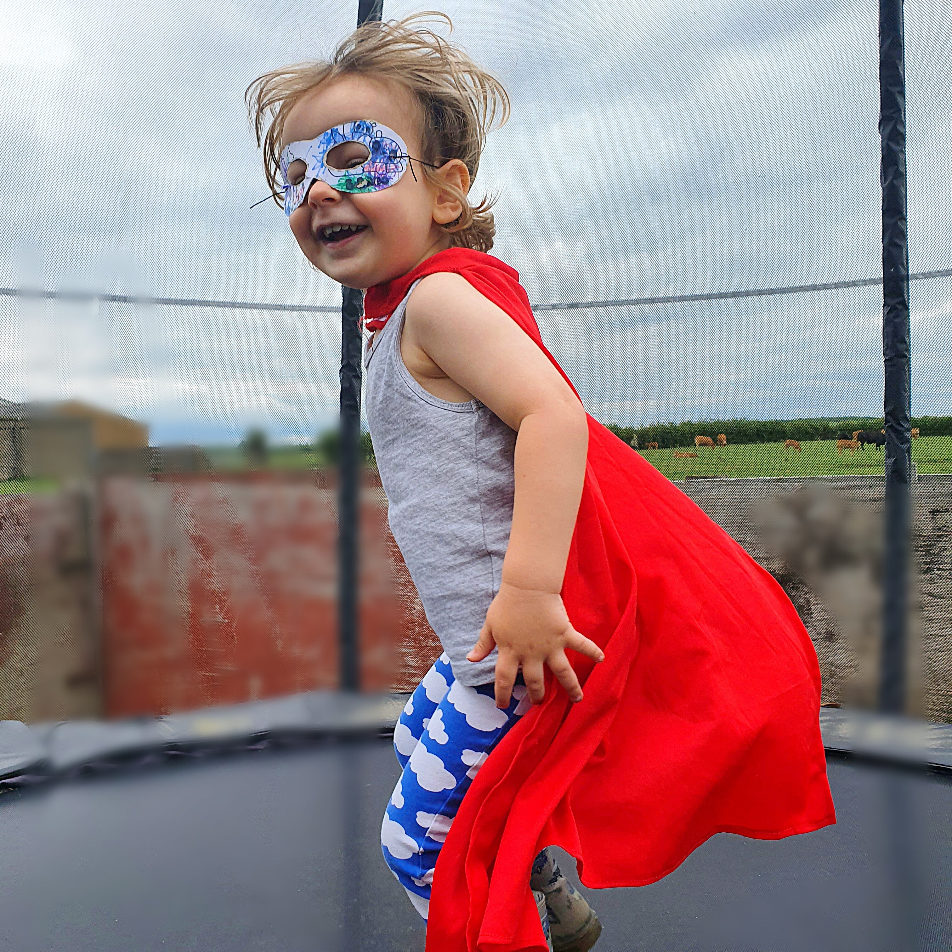 Happy child in a red cape and superhero mask jumping on a trampoline outdoors.