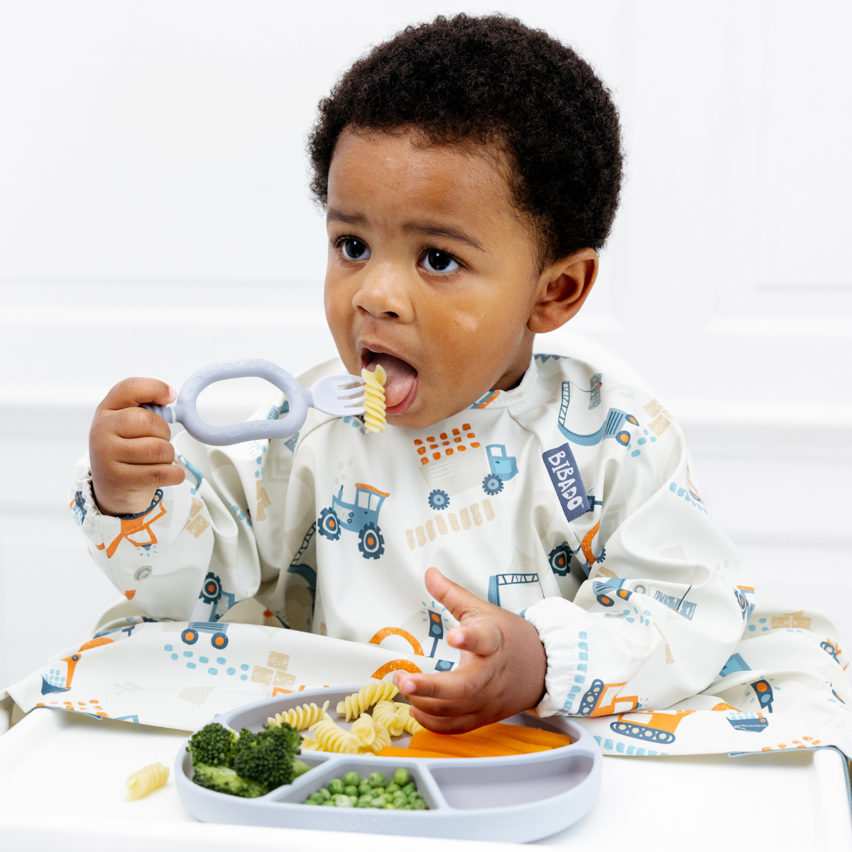 Toddler in a digger-print bib eating pasta and vegetables from a grey partitioned plate.
