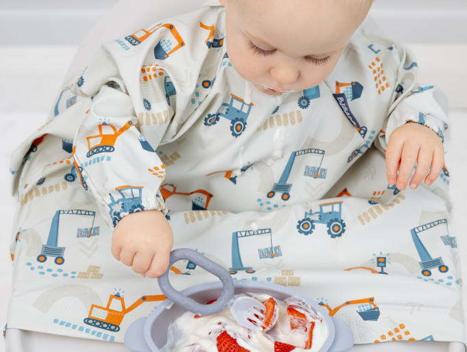Baby in a long-sleeved fabric bib with digger print, eating strawberries and yoghurt from a silicone bowl.