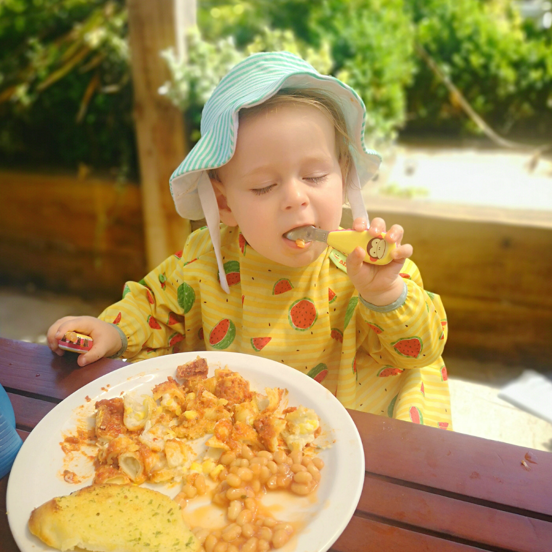 Toddler in a striped sun hat and watermelon bib eating baked beans and garlic bread outdoors.