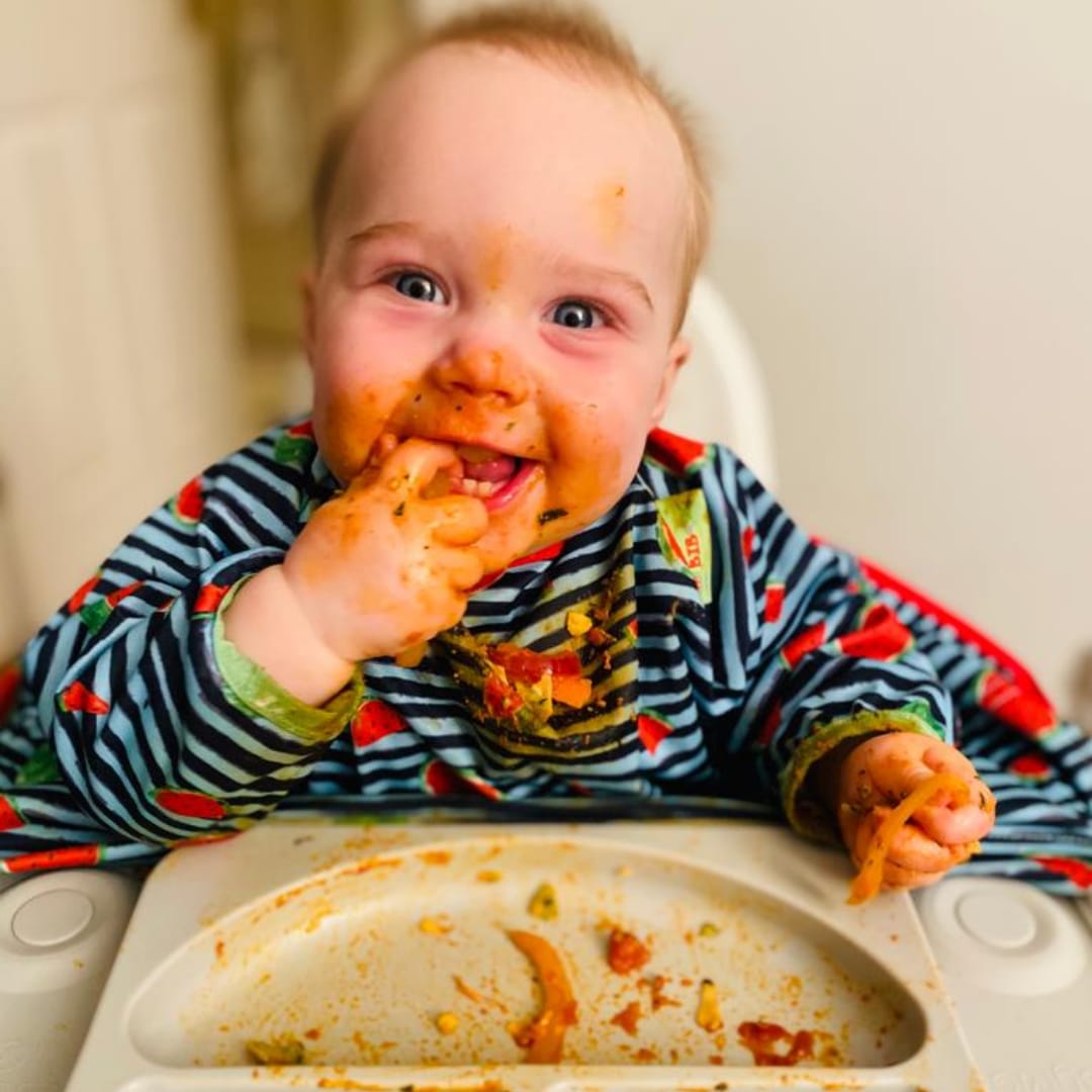 Happy baby in a highchair, covered in dairy-free weaning food, smiling brightly.
