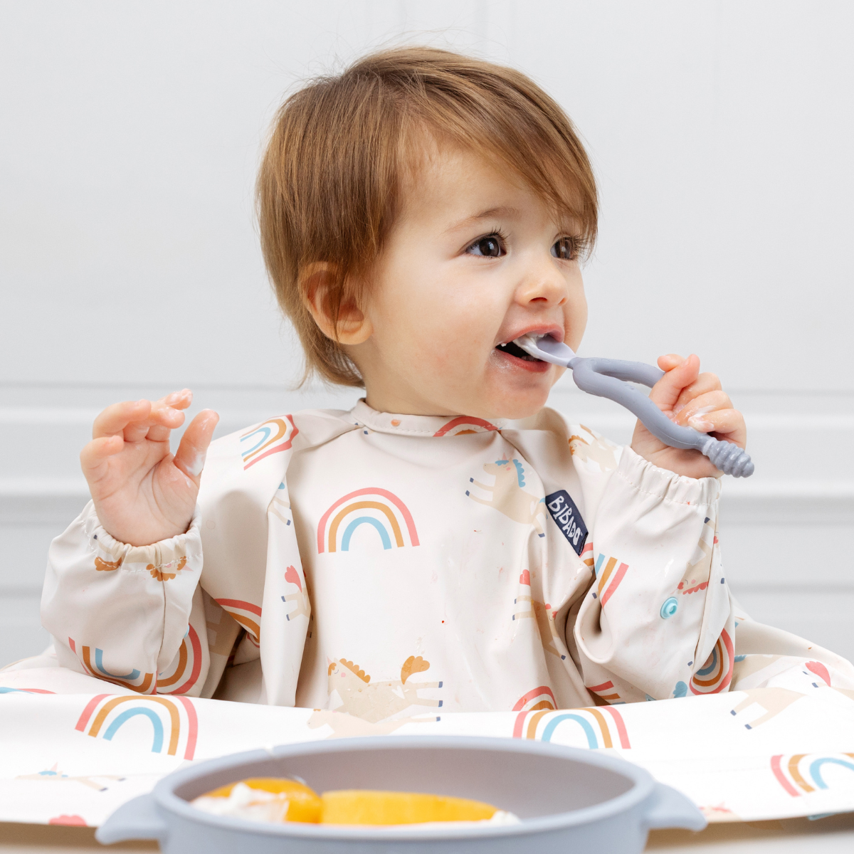 Toddler in a long-sleeved, rainbow-patterned coverall bib eating with a grey silicone spoon.