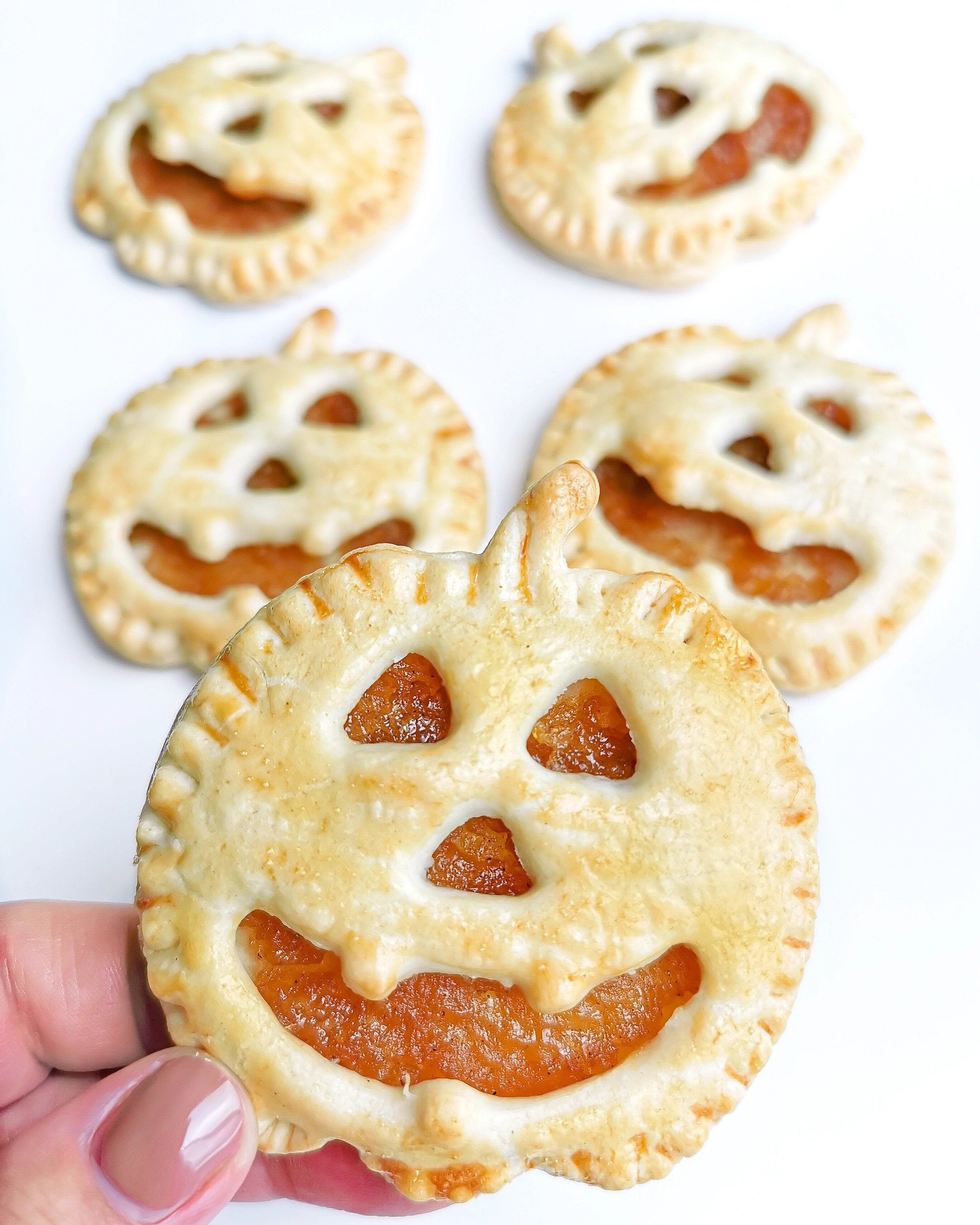 Hand holding a golden-brown pumpkin-shaped apple pie biscuit with a smiling jack-o'-lantern face.