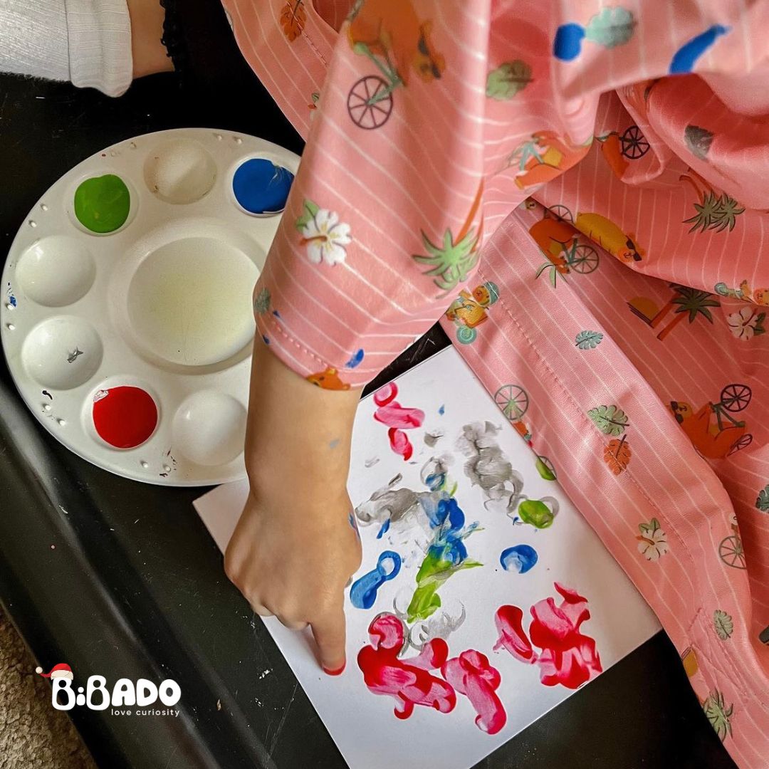 Child's finger painting colourful fingerprints on white paper next to a paint palette.
