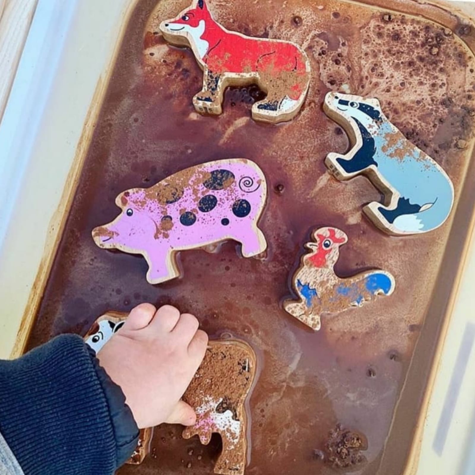 Child's hand playing with wooden farm animal toys in a muddy sensory tray.