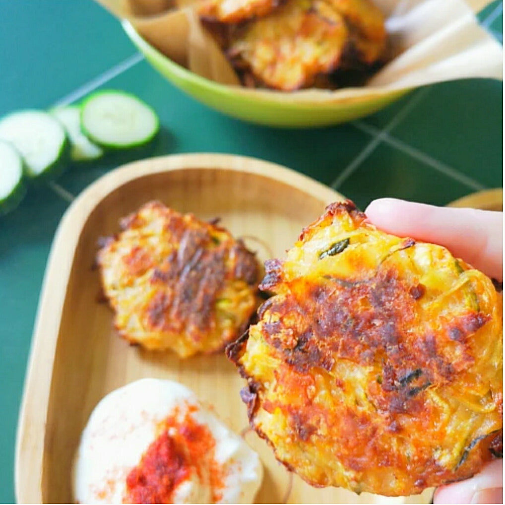 Hand holding a golden-brown courgette fritter from a wooden plate with dip and cucumber slices.