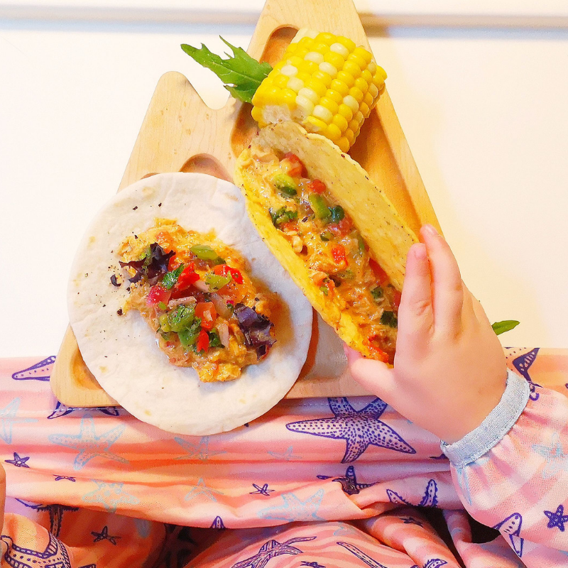 Child's hand reaching for a slow cooker chicken taco with corn on the cob on a wooden plate.