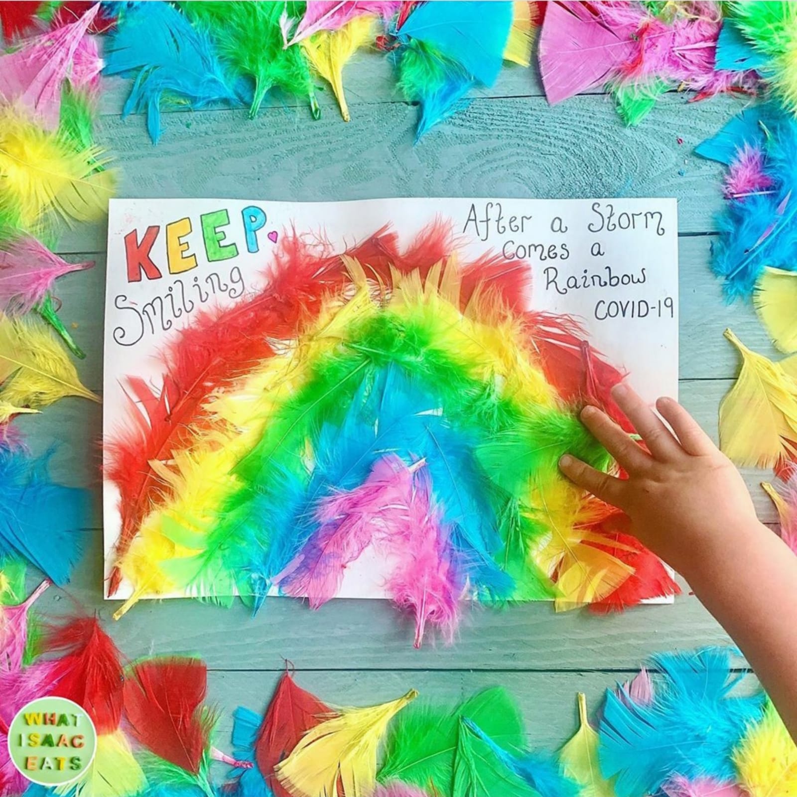 Child's hand arranging colourful feathers into a rainbow on paper, surrounded by loose feathers.