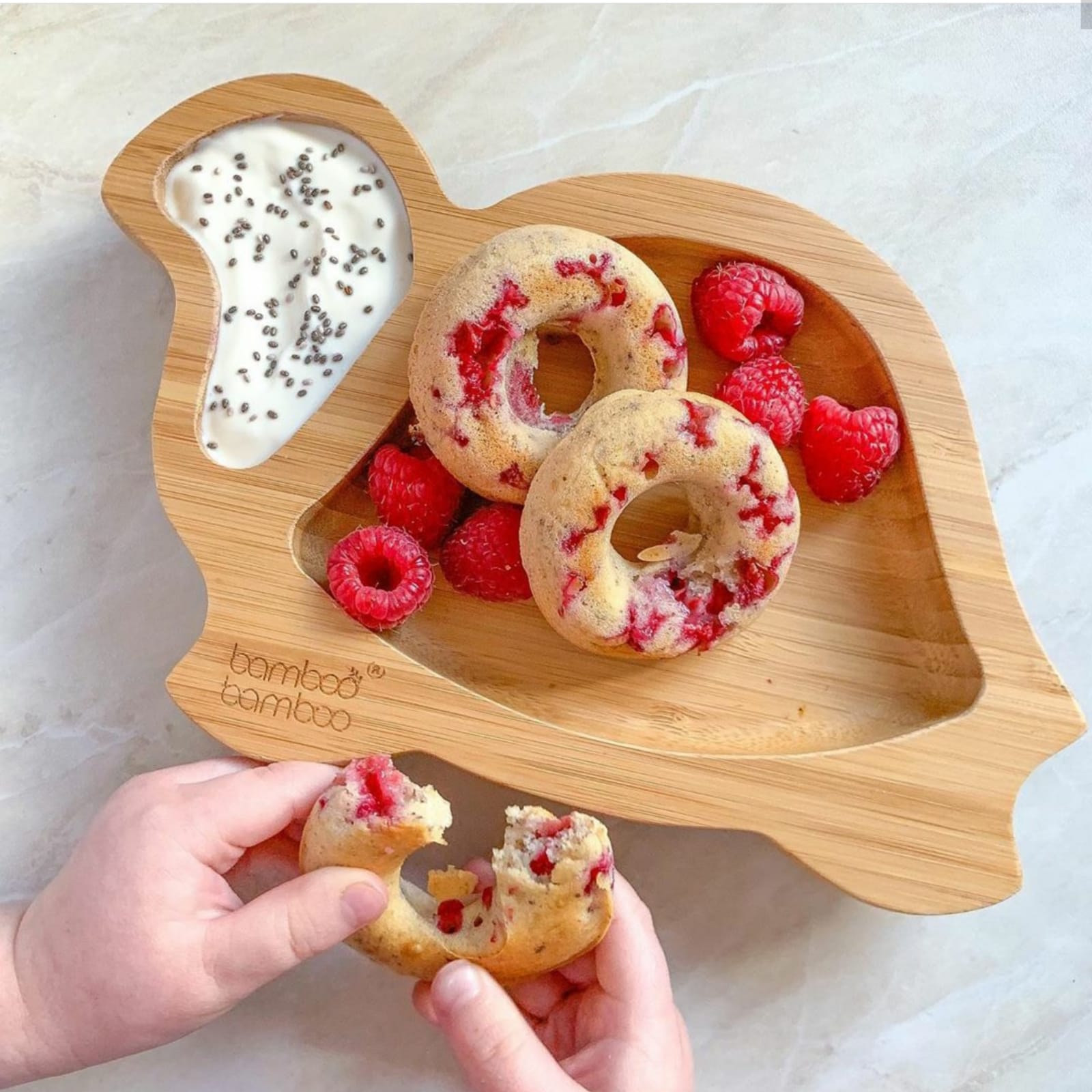 Child's hands holding a raspberry doughnut from a bamboo plate with more doughnuts, raspberries, and yoghurt.