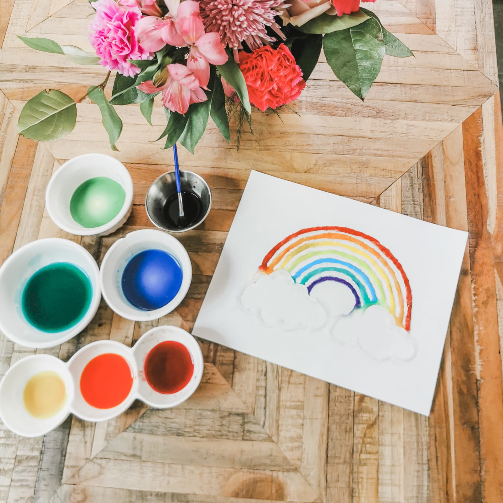 Salt painting of a rainbow with colourful paints, brushes, and flowers on a wooden table.
