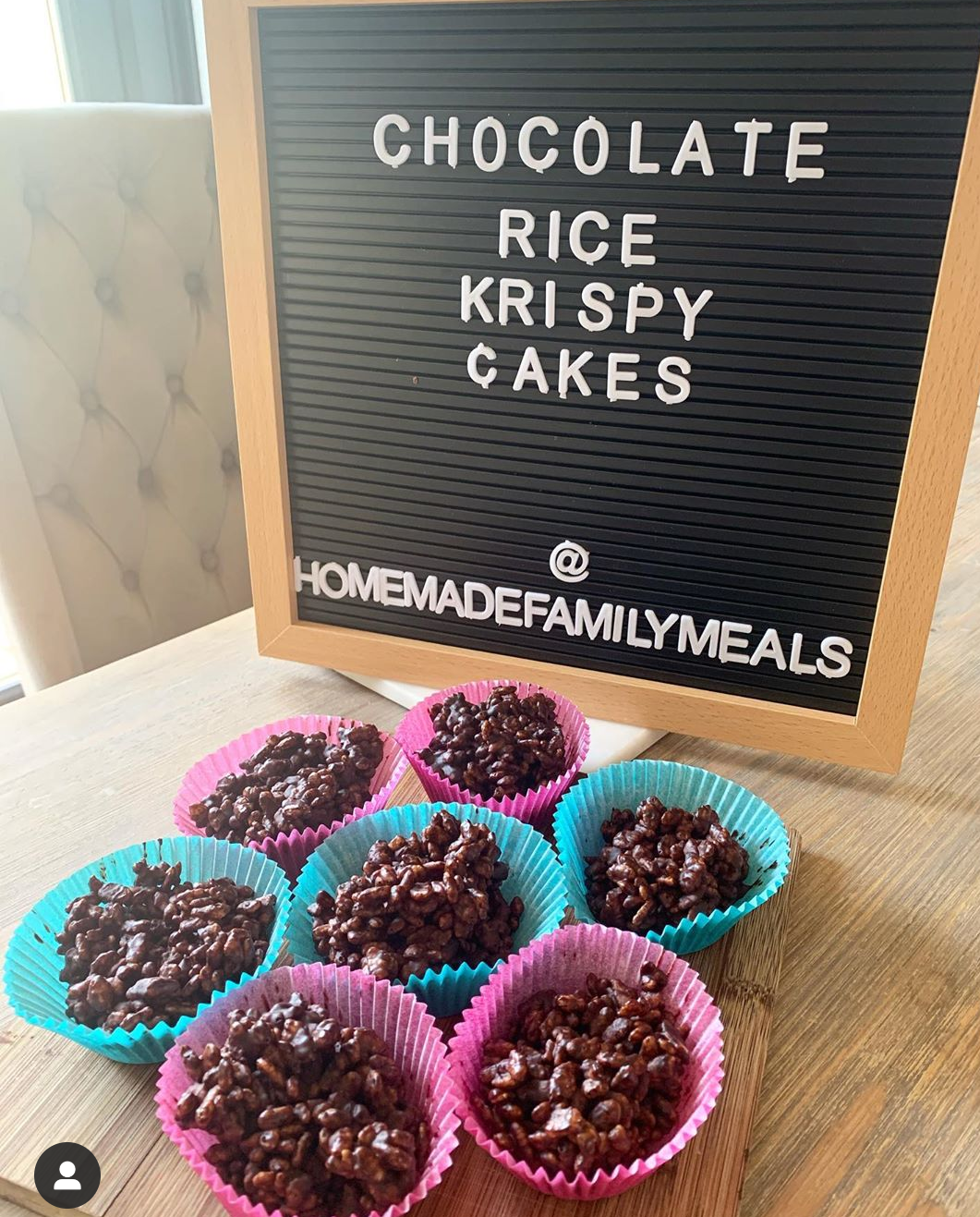 Seven chocolate Rice Krispie cakes in pink and blue cases, next to a letter board.
